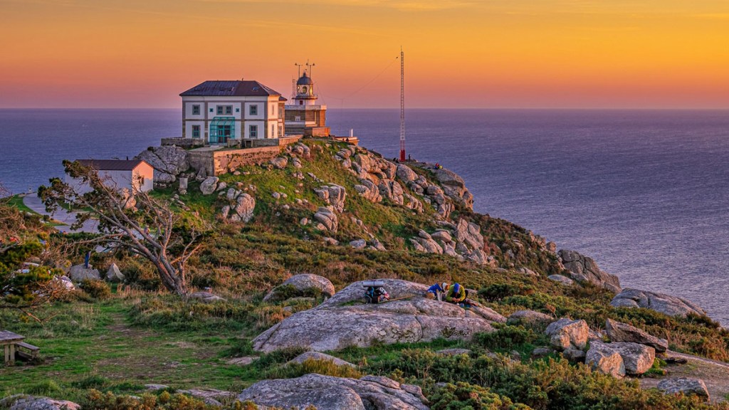 Ein Leuchtturm und ein historisches Gebäude auf einer felsigen Klippe an der Küste, umgeben von grünen Wiesen und Meer, bei Sonnenuntergang.
