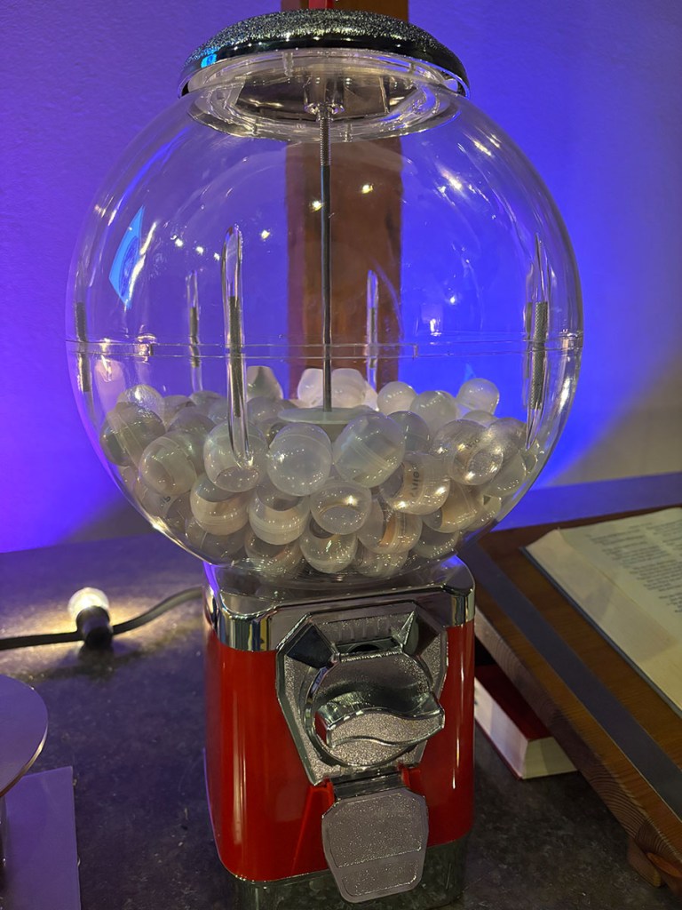Eine bunte Kaugummiautomat mit einer transparenten Kuppel, gefüllt mit runden Kaugummibällen, auf einem Tisch mit gedämpftem Licht.