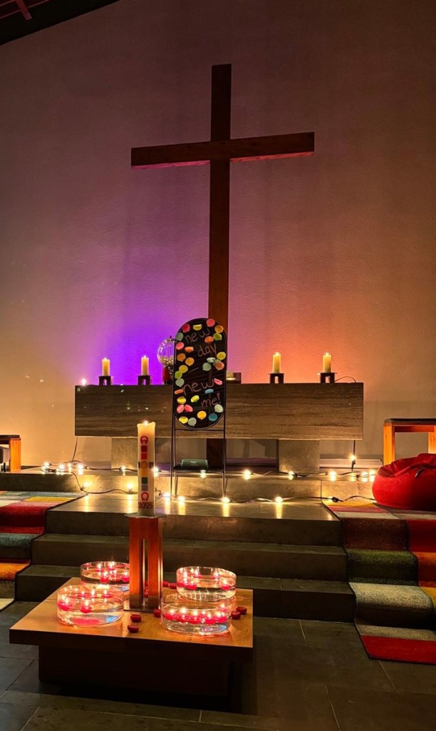 Ein dekorierter Kirchenraum mit einem großen Holzkreuz im Hintergrund, einem altartisch, Kerzen und einer Tischdekoration aus Wasserbecken mit roten Kerzen. Auf einem Schild steht 'neuer Tag'.
